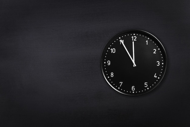 Фото Черные часы, показывающие без пяти минут до полуночи на фоне черной классной доски офисные часы, показывающие без пяти минут до полуночи или полудня