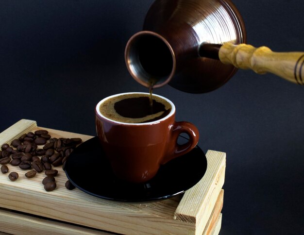 Black coffee in the brown cup on the wooden box  and turk for making coffee on the dark background. Close-up.