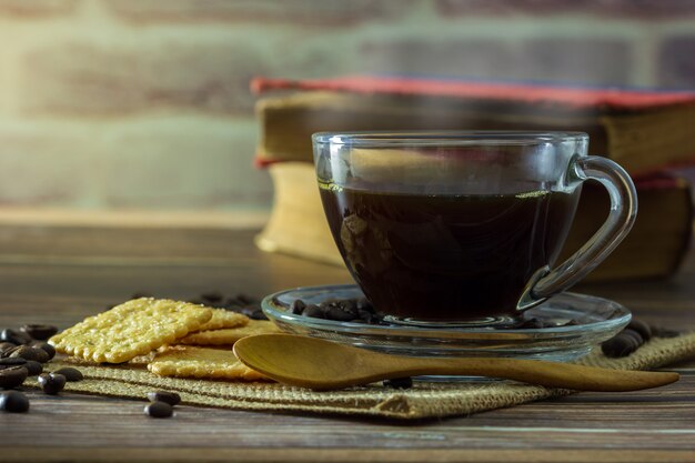 クリアガラスのカップとクラッカーと木製のテーブルの上の古い本とコーヒー豆のブラックコーヒー。