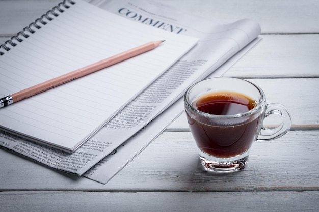 블랙 커피 컵, 공책, 신문 및 노트북이 테이블 위에 놓여 있습니다. 아침 뉴스, 아침