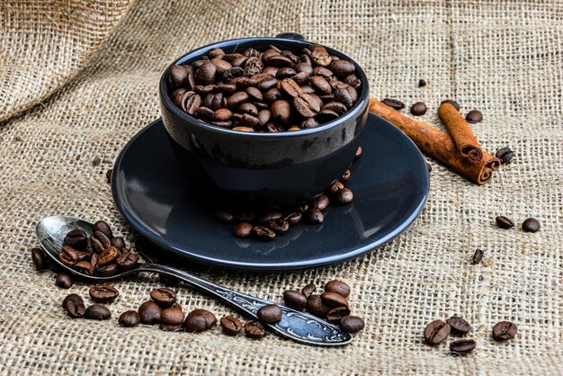 Foto una tazza di caffè nera piena di chicchi di caffè biologico e bastoncini di cannella su un panno di lino