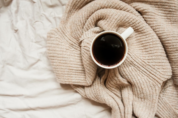 Black coffee in white mug in knitted beige sweater on rumpled bed top view monochrome cozy picture