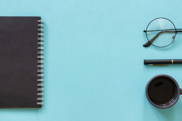 Black color notepad, cup of coffee, spectacles, pen on blue 