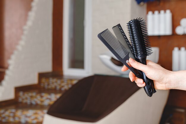 Foto pettini neri per capelli tagliati in mano femminile contro la sedia del lavandino del lavaggio dei capelli nello studio del salone di bellezza, interno del negozio di barbiere.