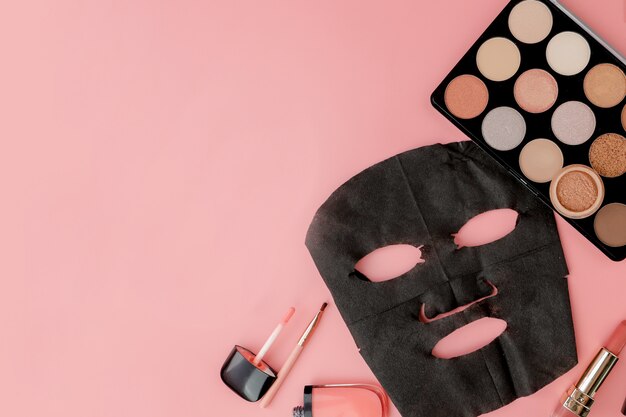 Black cosmetic mask and makeup set on pink background with copy space