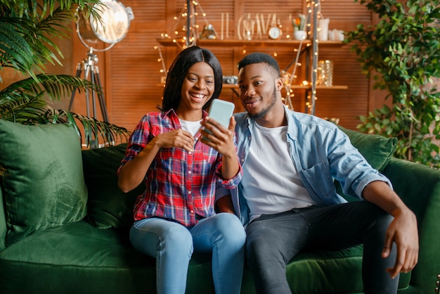 Black couple sitting on sofa and looking on mobile phone at home. Happy african love couple leisures in their house, cheerful family relaxing together