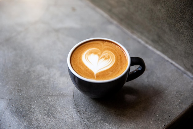 Black cup of cappuccino with latte art of heart shape on saucer on concrete background