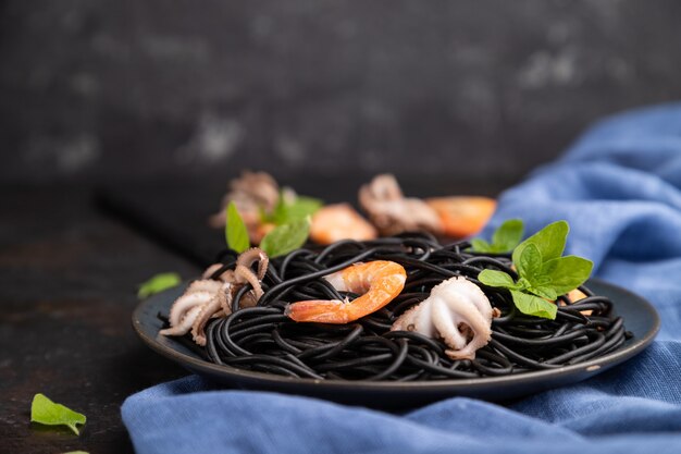 Black cuttlefish ink pasta with shrimps or prawns and small octopuses on black concrete background and blue textile. Side view, close up, selective focus.