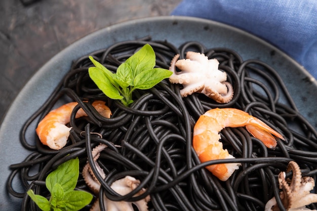 Black cuttlefish ink pasta with shrimps or prawns and small octopuses on black concrete background and blue textile. Top view, close up, selective focus.