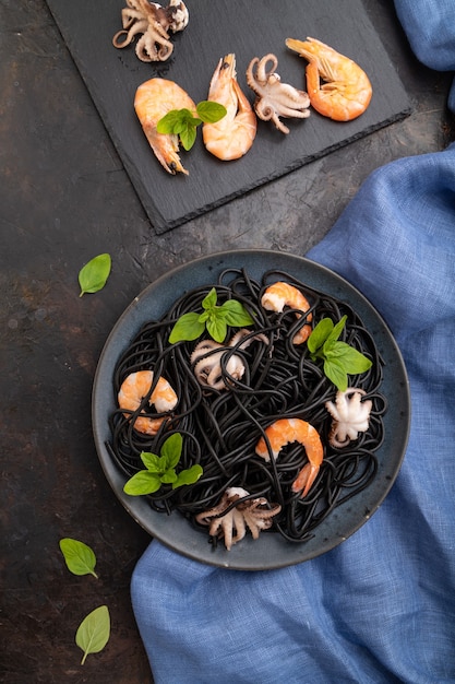 Black cuttlefish ink pasta with shrimps or prawns and small octopuses on black concrete background and blue textile. Top view, flat lay, close up.