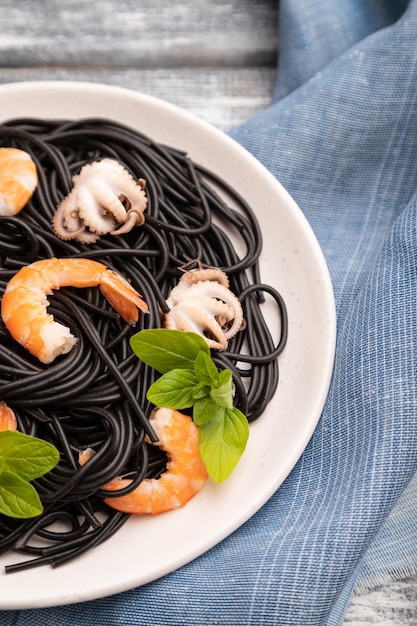 Black cuttlefish ink pasta with shrimps or prawns and small octopuses on gray wooden background and blue textile. Side view, close up, selective focus.