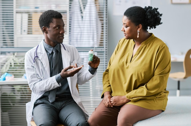 Black doctor holding bottle of pills consulting woman in clinic