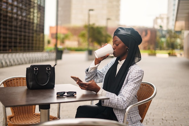 Donna nera elegante che beve caffè e naviga nello smartphone