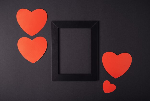 Black empty photo frame with red valentine hearts on the dark (black) background. Valentine's Day concept. Flat lay, top view. Blank space for text.