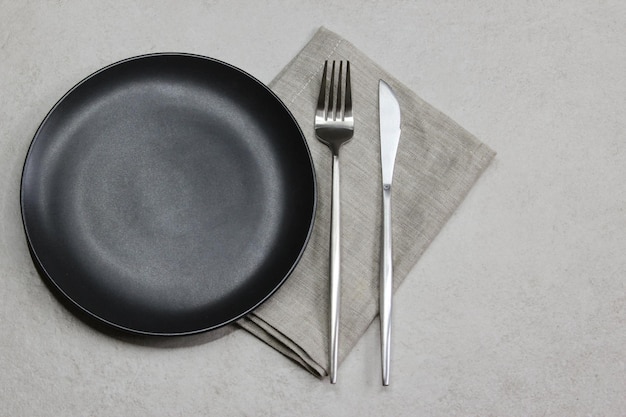 Черная пустая тарелка с ножом и вилкой с серой тканевой салфеткой на сером каменном фоне