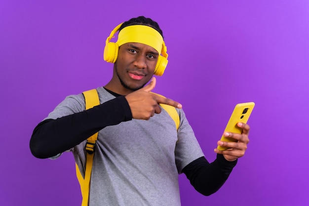 배낭과 보라색 배경 학생 개념에 노란색 헤드폰을 가진 흑인 민족 남자가 전화를 가리키는