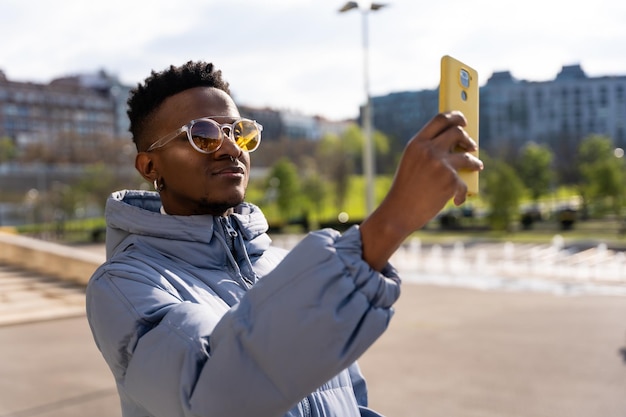 A black ethnic man with a phone and a blue jacket in the city taking a selfie