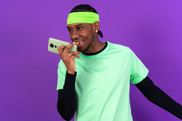 Черный этнический мужчина с телефоном в зеленой одежде, изолированный на фиолетовом фоне, улыбается голосовым сообщением