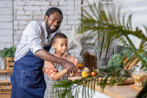 흑인 아버지와 그의 아들 아이가 집에서 부엌에서 신선한 야채를 요리하고 있습니다. 아프리카 아빠와 소년 아이가 함께 준비합니다
