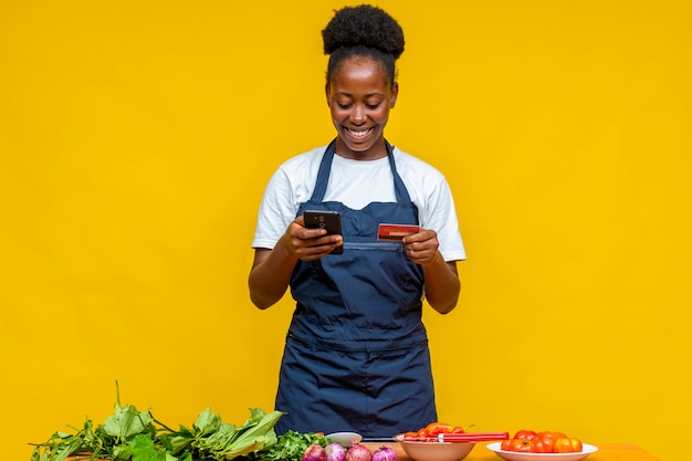 Black female cook using her phone and credit card, with vegetables and other farm produce in front of her