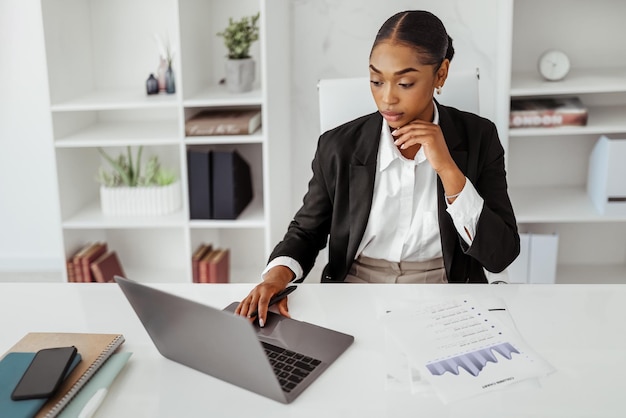 Чернокожая женщина-предпринимательница работает с ноутбуком и бумагами на рабочем месте в офисе, управляя финансами