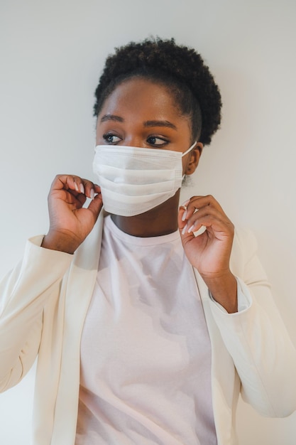 Фото Черная женщина с вьющимися волосами надевает медицинскую маску, стоя в комнате с закрытым воздухом и глядя в сторону изолированная над белой стеной респираторный вирус медицинское здоровье портрет лица молодой женщины