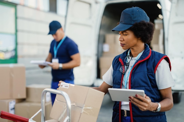 Black female worker using digital tablet while doing package inventory before the delivery Her coworker is in the background