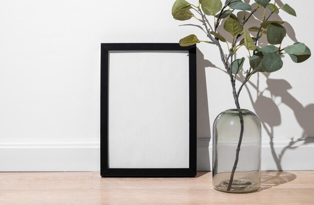 A black frame with a blank canvas against a light wall and on a wooden floor with vase and branch