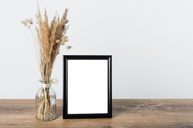 Photo next to the black frame on a wooden table is a glass vase with dried herbs. light background.