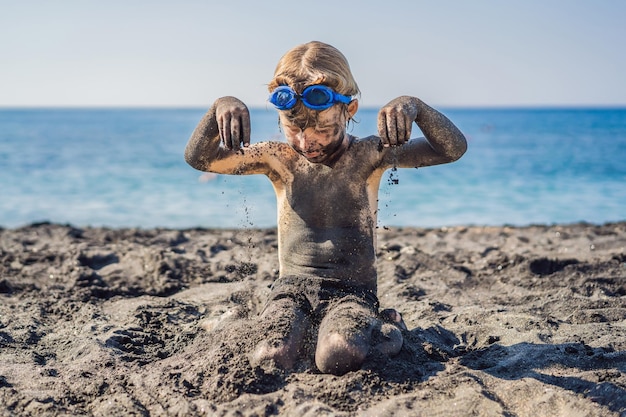 Black friday concept smiling boy with dirty black face sitting\
and playing on black sand sea beach