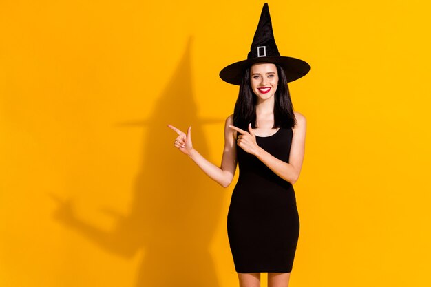 블랙프라이데이 할인. 귀여운 매력적인 젊은 마녀 마술사 직접 손가락 빈 공간 빛나는 미소의 사진은 밝은 노란색 배경에 고립 된 검은 모자 드레스를 입고