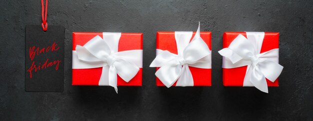 Black Friday - handwritten inscription on the tag. Red gift boxes with white ribbons. Black concrete background.