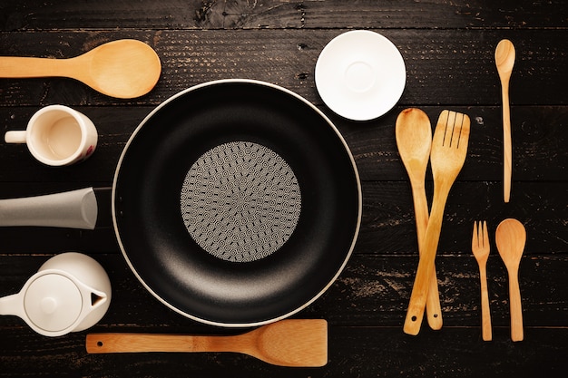 Black frying pan with cooking utensils on black wood table background.