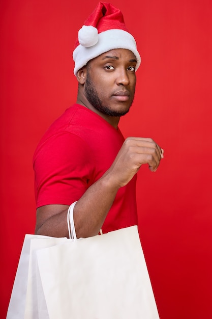 Black gay man in a Santa hat looks at the camera and holds white paper bags in his hands