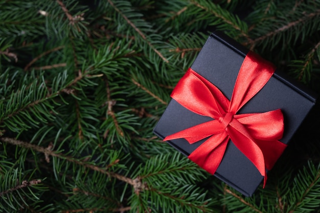 Black gift box with red ribbon bow on full frame fir branches background. Merry christmas card. High quality photo