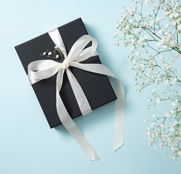 A black gift box with a white ribbon on a blue background with a bouquet of  gypsophila. The concept of a holiday or gift card. Top  view and copy space.