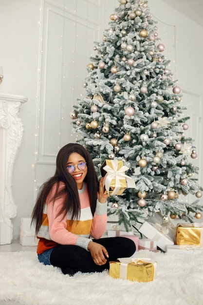 Black girl in eyeglasses near Christmas tree opening gift at home