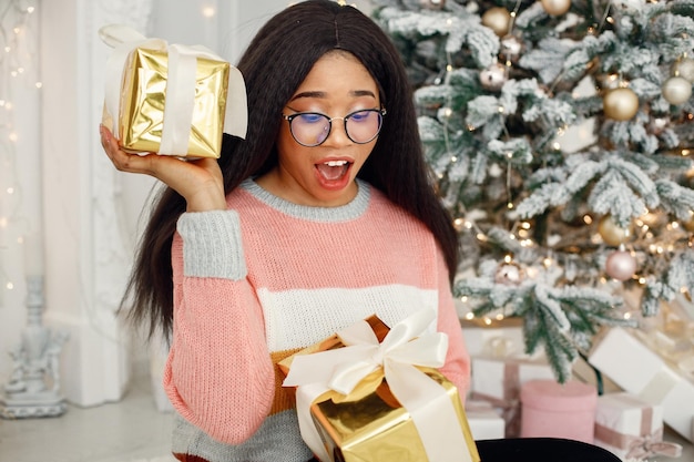 Black girl in eyeglasses near Christmas tree opening gift at home