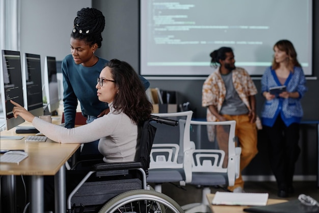 Photo black girl and it specialist in wheelchair writing code together in office