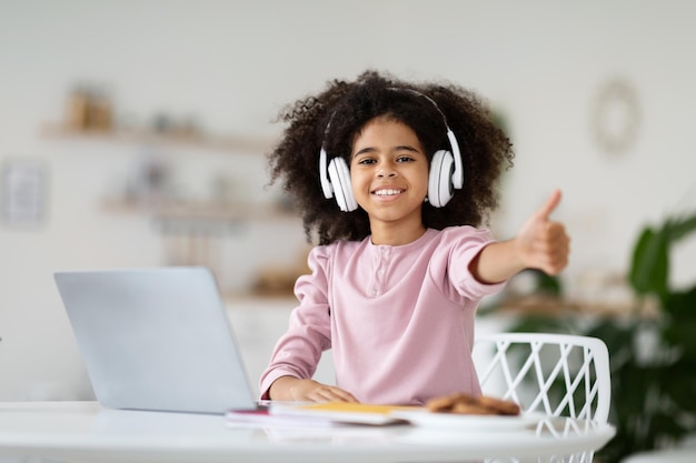 Scolara ragazza nera che utilizza laptop e cuffie che mostrano il pollice in su