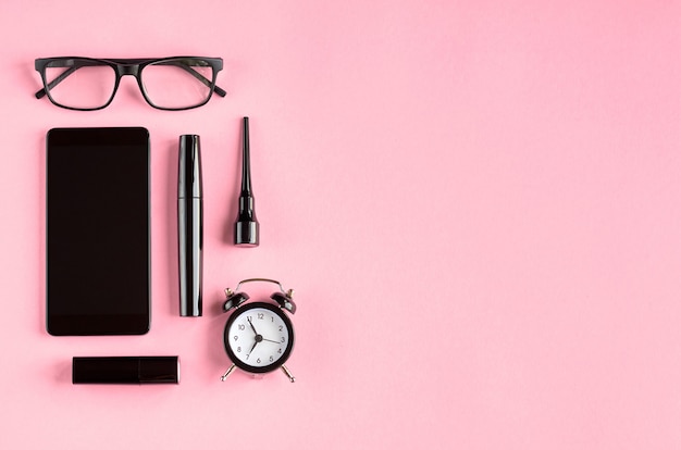 Black glasses, alarm clock, cellphone, mascara and pomade on pink surface.