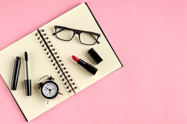 Photo black glasses, alarm clock, paper notepad, mascara and red pomade on pink surface.