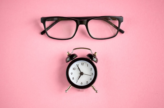 Black glasses and alarm clock on pink surface.