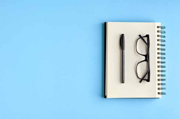 Black glasses, pen and paper notebook on blue surface.