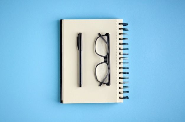 Black glasses, pen and paper notebook on blue surface.