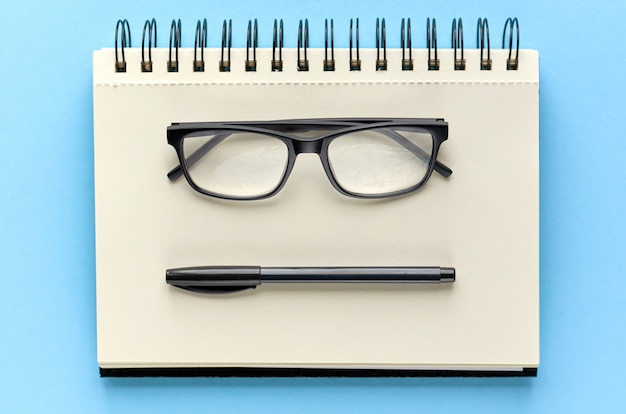 Black glasses, pen and paper notebook on blue surface.