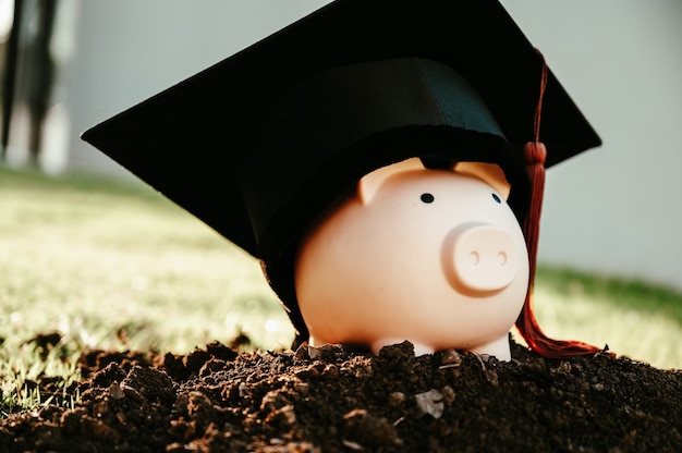 A black graduate hat, brown tassels and a pink piggy bank lay on the black ground.