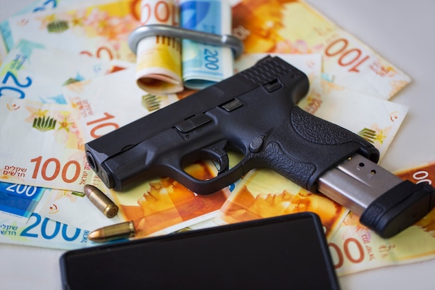 Black gun pistol with ammunition and mobile phone on on stack money Israeli New Shekels banknotes on table. Semi Automatic handgun firearm with New Israel Shekel 100, 200 NIS bills. Currency, criminal