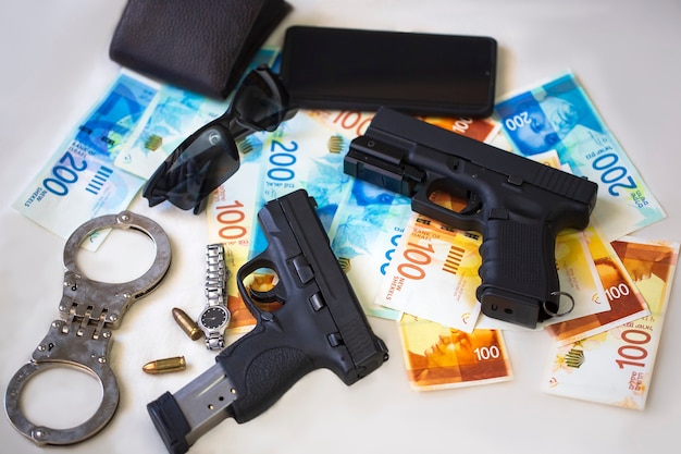 Black guns pistols with ammunition, handcuffs, sunglasses,\
wrist watch and mobile phone on on money israeli new shekels\
banknotes on table. semi automatic handgun firearm with new israel\
shekel bills