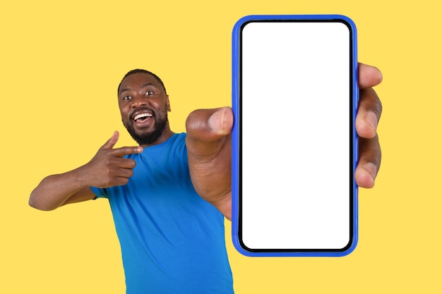 Черный парень показывает смартфон с большим пустым экраном на желтом фоне